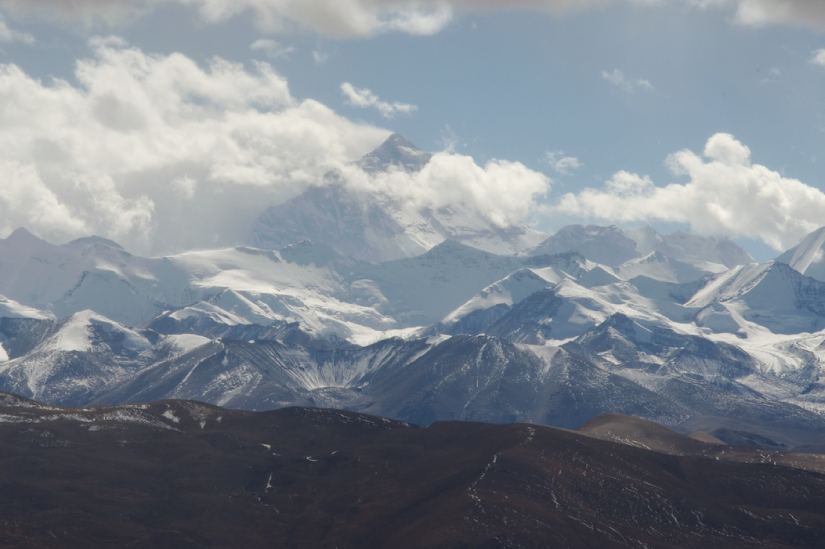 Ljósmyndin sýnir Everestfjall úr fjarska. Fjallið er stórt, pýramídalaga og klettótt og í forgrunni eru margir minni, snævi þaktir tindar. Ský hylja tind Everestfjalls að hluta.