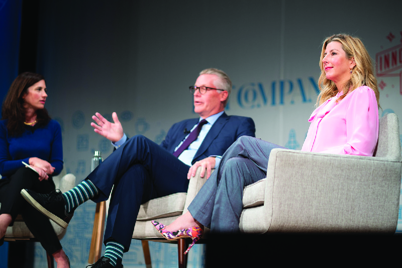 Ljósmynd af þremur einstaklingum að ræða saman á sviði; Sara Blakely situr til hægri.
