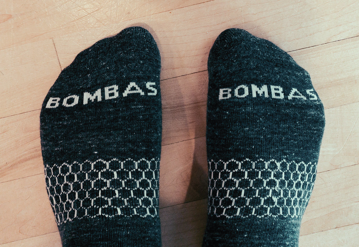 Par af dökkgráum Bombas-sokkum með hvítu hunangskökumynstri, á fótum sem hvíla á ljósu viðargólfi.