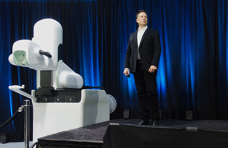 Elon Musk stendur á sviði við stóra og straumlínulaga vél með sléttum flötum og ávölum hornum.