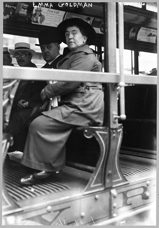 Ljósmynd sýnir Emmu Goldman sitja á bekk í sporvagni með tveimur körlum.