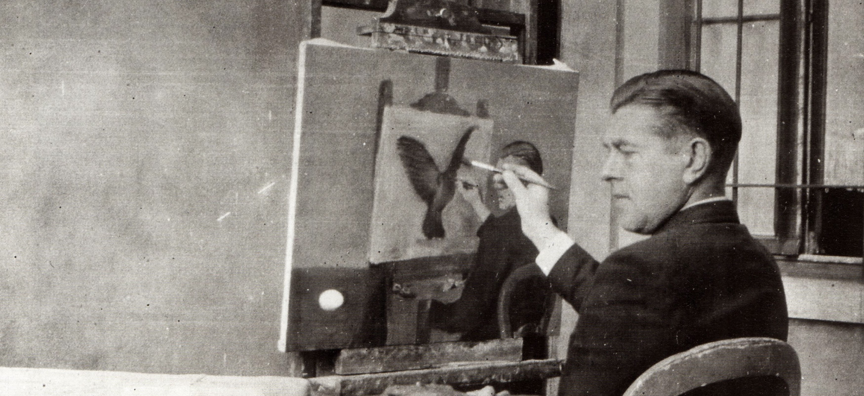 Svarthvít ljósmynd af René Magritte með pensil við trönur og nær fullgert málverk