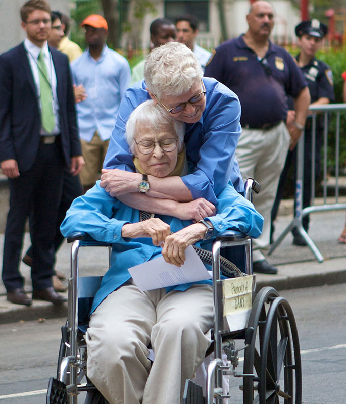 Phyllis Siegel og Connie Kopolev faðmast á götu í New York-borg á meðan fólk fylgist með. Kopolev situr í hjólastól og heldur á skjali.