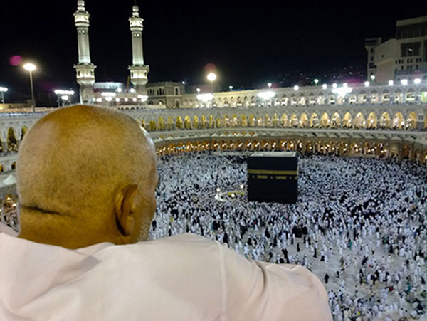 Maður í hvítum klæðum sést aftan frá horfa yfir Kaaba, helgasta stað íslams. Hundruð annarra, öll í svörtum eða hvítum klæðum, ganga í hring um stóra svarta teningslaga byggingu á gólfi mannvirkis sem líkist leikvangi.