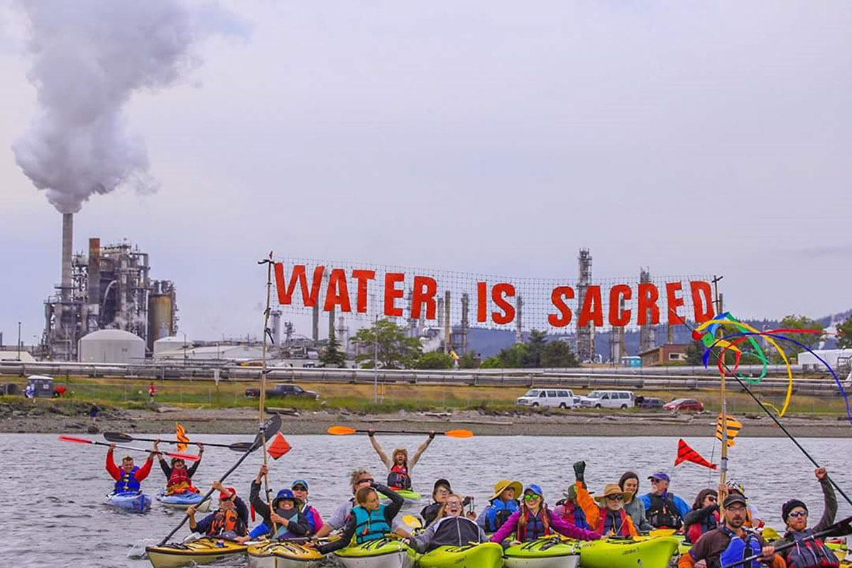 Um tuttugu manns á kajökum fljóta fyrir framan olíuhreinsistöð. Þau halda á skilti með ensku áletruninni „Water Is Sacred“ („Vatn er heilagt“).