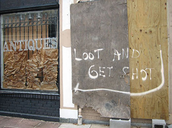 Ljósmynd af skemmdu húsi eftir fellibylinn Katrínu. Á girðingunni er skilti með ensku áletruninni „Looters Will Be Shot“ („Skotið verður á ræningja“).