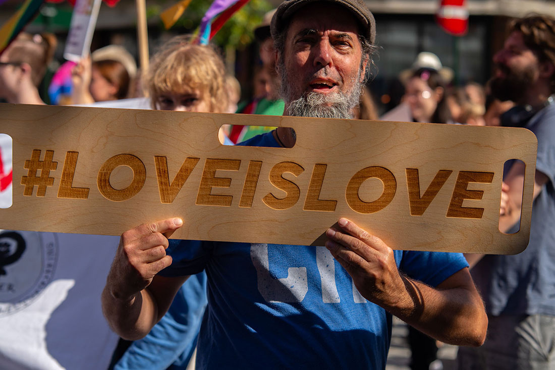 Manneskja heldur á skilti í göngu eða á samkomu. Á skiltinu er myllumerki og orðin „Love Is Love“.