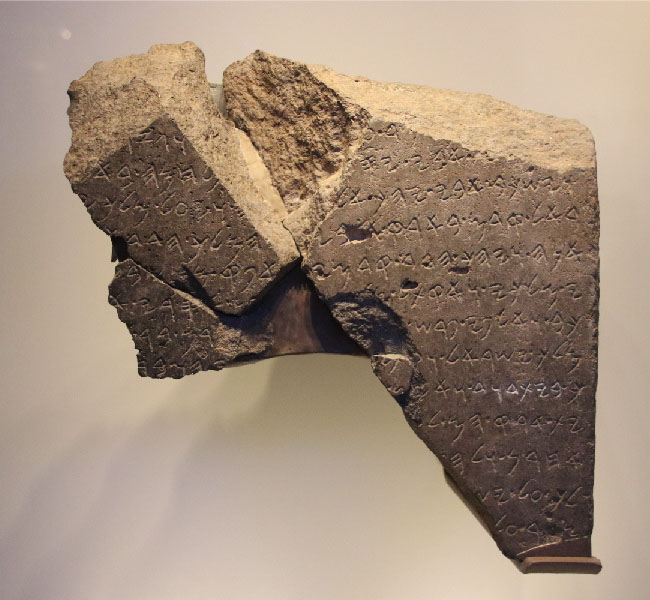 Sýnt er brot úr brúnleitum steini. Áletranir sjást um allan steininn.