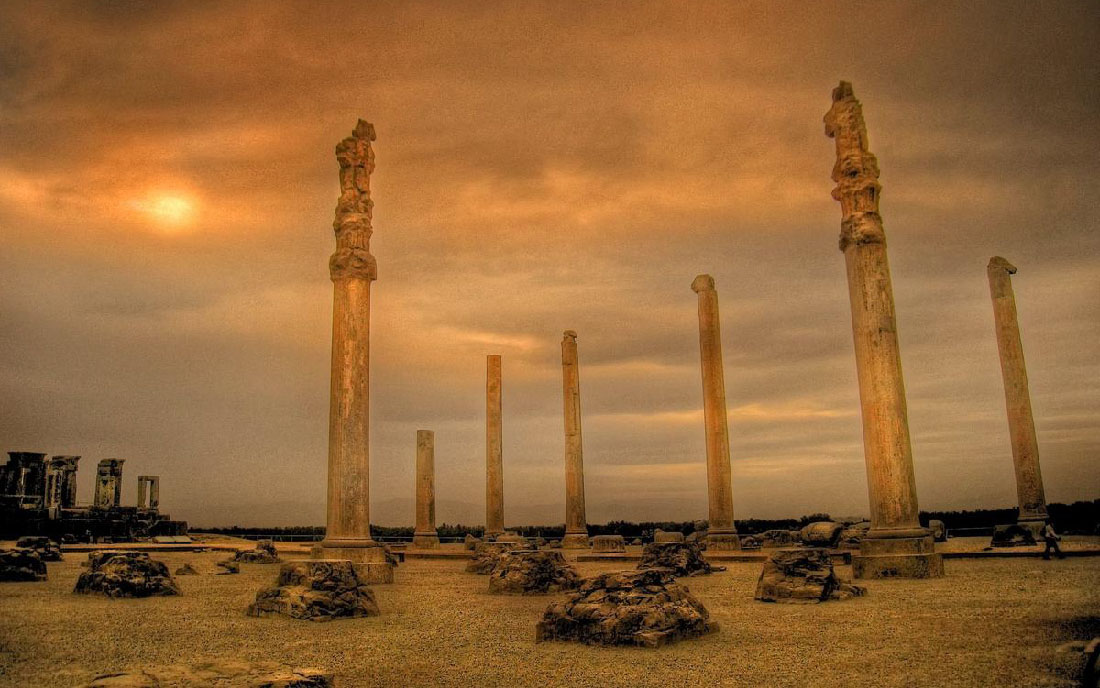 Á myndinni sjást sjö brotnar, brúnleitar súlur við sólarlag. Sumar súlnanna eru með skrautverkum efst. Undirstöður liggja á víð og dreif um jörðina. Vinstra megin í bakgrunni eru brotnar tvöfaldar súlur ofan á stórum, dökkum palli. Jörðin er malarborin og tré sjást langt í burtu.