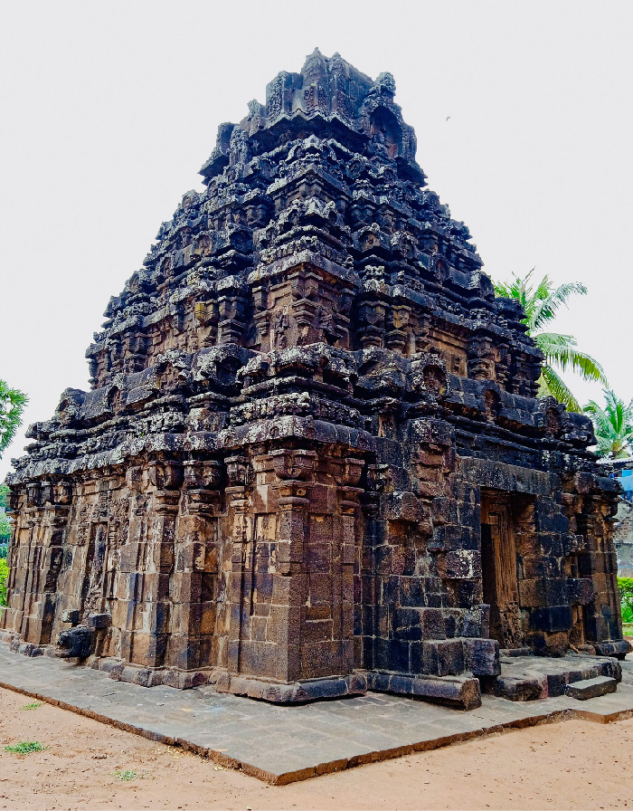 Mynd sýnir þrepaskipta, píramídalaga steinbyggingu. Hún er brún, blá og svört, með dyrum hægra megin og tveggja þrepa tröppum. Engir gluggar eða önnur op sjást. Pálmatré gægjast fram undan byggingunni í bakgrunni og ljósbrúnn moldarflötur umlykur gráan, flísalagðan grunn hennar.