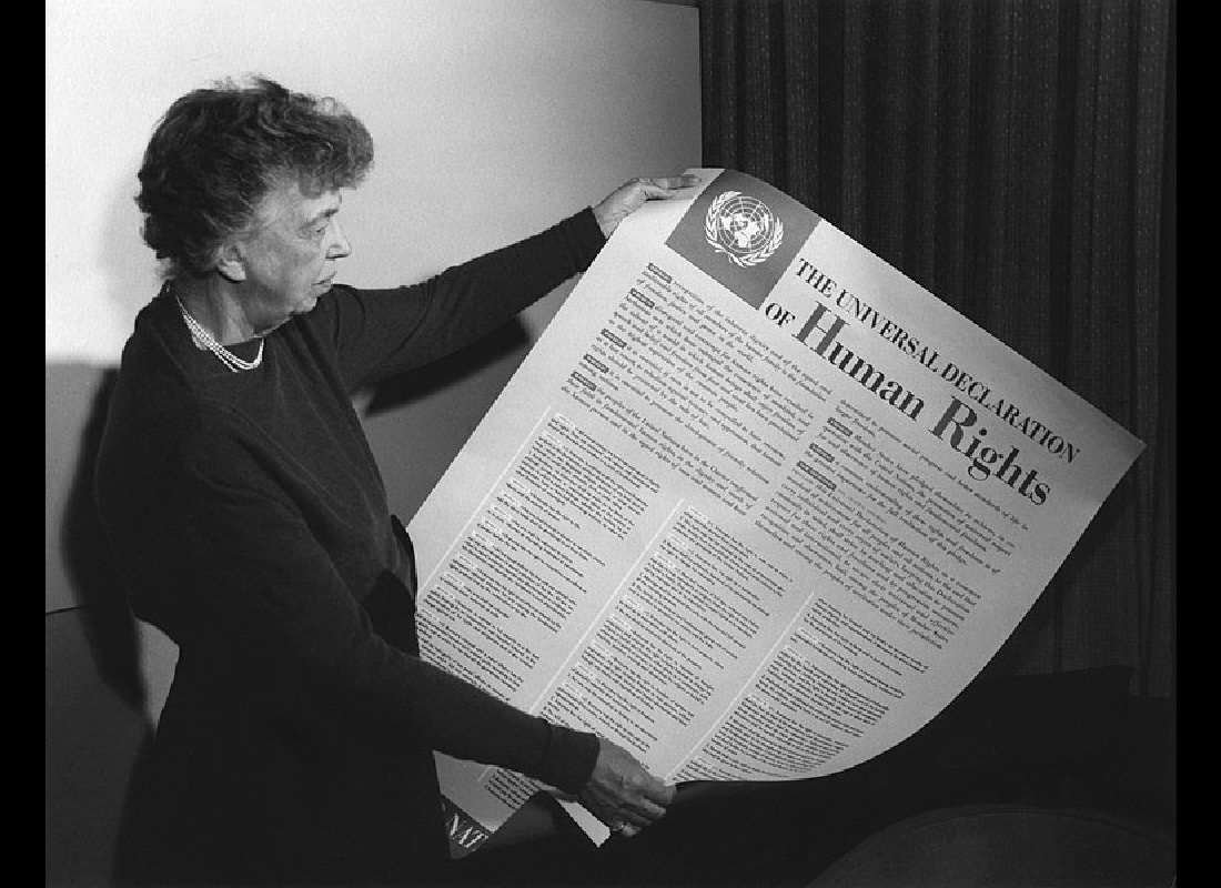 Ljósmyndin sýnir Eleanor Roosevelt halda á stóru skjali með yfirskriftinni „Mannréttindayfirlýsing Sameinuðu þjóðanna“. Merki Sameinuðu þjóðanna er efst í vinstra horni skjalsins.
