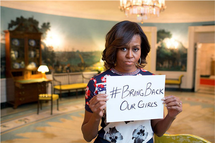 Mynd sýnir bandaríska konu með dökkt hörund standa og halda á litlu hvítu blaði með áletruninni „#Bring Back Our Girls“ með svörtu bleki. Hún er með stutt liðað hár, stóra hringeyrnalokka, bláan, rauðan og hvítan blómakjól og demantshring á vinstri hendi. Í bakgrunni sést óskýrt herbergi með háu skrifborði, stól, bekkjum og lampa og landslagsmálverk skreyta veggina. Stór ljósakróna úr gleri hangir yfir höfði hennar í bakgrunni.