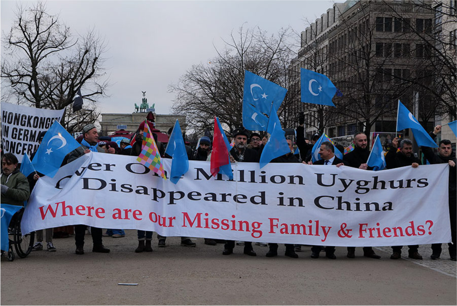 Mynd sýnir fólk bera stóran hvítan borða á götu fyrir framan stórt minnismerki með grænni styttu efst, tré og háa byggingu til hægri. Á borðanum stendur „Over One Million Uyghurs Disappeared in China“ með svörtu og síðan „Where are out Missing Family & Friends?“ með rauðu neðst. Nokkrir einstaklingar í jakkafötum, kápum og með hatta og trefla halda á borðanum. Þeir bera einnig fána. Fánarnir eru flestir bláir með tungli og stjörnu, þrír eru köflóttir í ýmsum pastellitum og einn rauður. Vinstra megin við borðann heldur maður í hjólastól á bláum fána. Aftar til vinstri er hvítur borði með orðunum „Hongkingers in Germa Concern“ en sumir stafirnir eru huldir öðrum fánum.